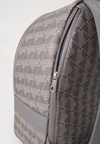 Zaino grigio con un motivo geometrico in rilievo. Presenta una zip liscia, un tessuto resistente e dettagli simili al cuoio. Forma compatta.