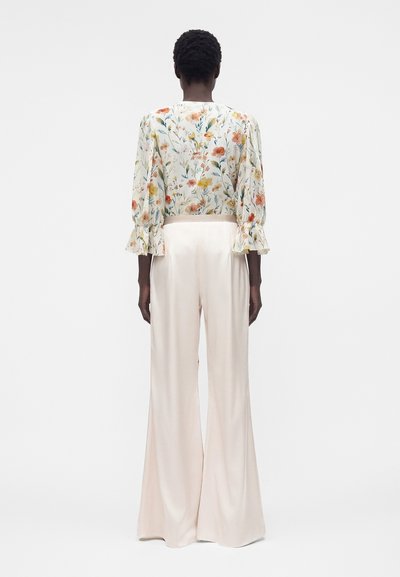 Άτομο με κοντά, σγουρά μαλλιά που φοράει μπλούζα με λουλούδια μακρυμάνικη και μπεζ παντελόνια με ψηλή μέση και φαρδιά πόδια, στέκεται γυρισμένο με την πλάτη.