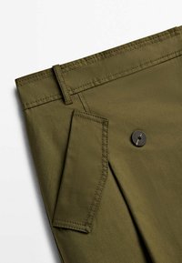 Gros plan sur un pantalon vert olive montrant une boucle de ceinture, une poche à rabat avec des coutures et un seul bouton sombre sur le tissu.
