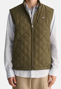 Olive green, quilted vest with a high collar and zip closure, featuring side pockets and a small emblem on the chest. Worn over a striped shirt.