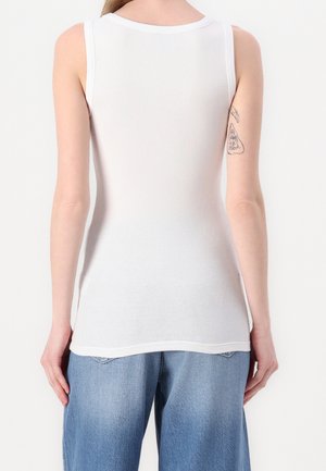 Vrouw draagt een mouwloos wit geribd tanktopje en losse blauwe jeans, en toont een kleine tatoeage op de achterkant van haar bovenste linkerarm.