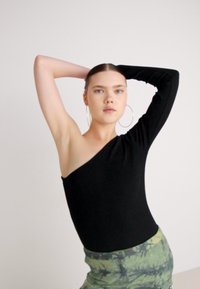 Crni bodysuit s jednim ramenom napravljen od rastezljivog materijala; model pozira s podignutim rukama, noseći velike srebrne naušnice u obliku kruga i suknju s zelenim uzorkom.