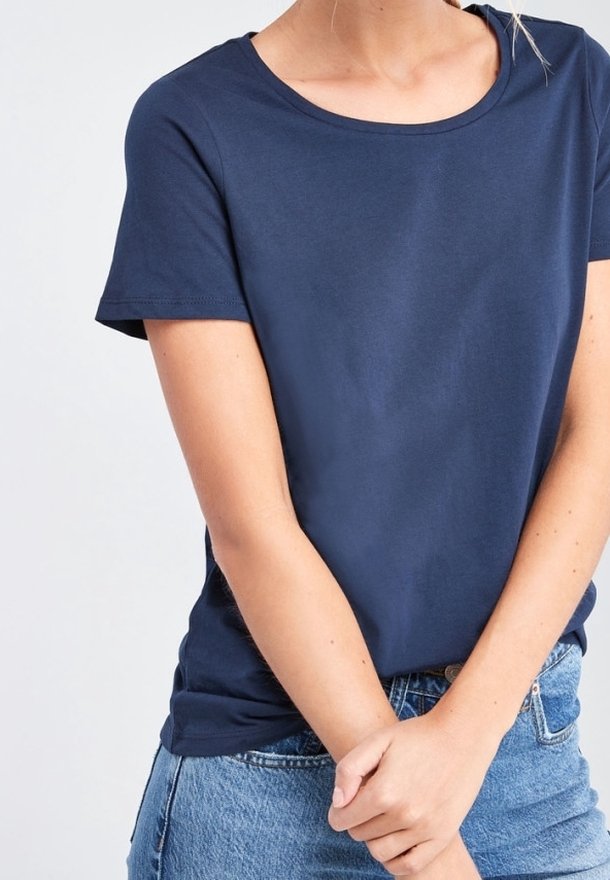 Personne portant un t-shirt uni bleu marine à manches courtes et un jean bleu, les bras croisés à la taille, sur un fond blanc.