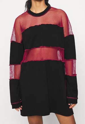 Vrouw draagt een zwarte jurk met horizontale donkerroze visnetpanelen op de borst, mouwen en taille.