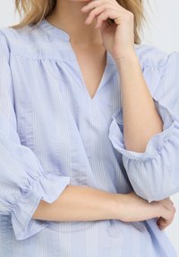 Femme portant un chemisier bleu clair rayé à manches longues avec un col en V, reposant un bras sur son corps et touchant son menton avec la main.