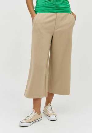 Pantalons larges beige avec poches, en tissu doux. Portés avec des baskets beiges. Un top vert froncé complète la tenue.
