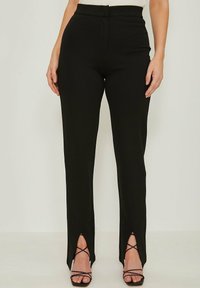 Mujer vestida con pantalones negros de cintura alta con aberturas en la parte inferior, combinados con tacones negros de tiras, sobre un fondo sencillo.