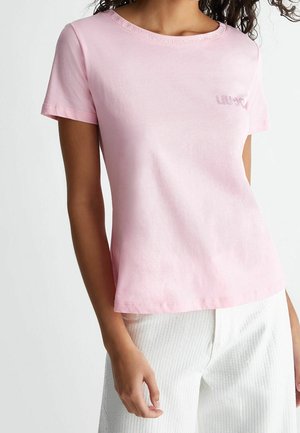 Frau trägt ein hellrosa kurzärmeliges T-Shirt mit Schriftzug am Kragen und weiße, strukturierte Hose vor weißem Hintergrund.