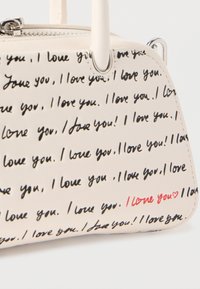 Κρεμ τσάντα χειρός με μαύρο χειρόγραφο κείμενο «Σ' αγαπώ» επαναλαμβανόμενο, ένα κόκκινο «Σ' αγαπώ» με καρδιά, και ασημένιο φερμουάρ και κρίκους λαβής.