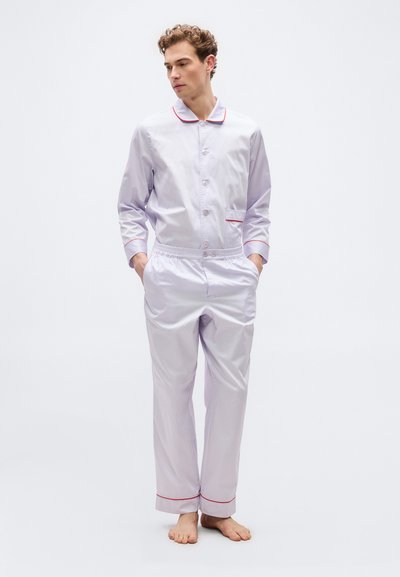 Νέος άνδρας σε ανοιχτόγριες πυτζάμες με κόκκινη λεπτομέρεια, όρθιος ξυπόλητος, με τα χέρια στις τσέπες, κοιτάζοντας προς τα πλάγια μπροστά σε απλό φόντο.