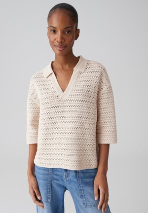 Femme aux cheveux tirés porte un pull en maille beige clair à encolure en V avec col et manches trois-quarts, associé à un jean en denim bleu, debout devant un fond gris.