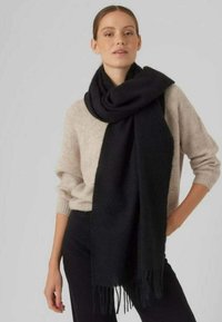 Svart skjerf laget av mykt materiale, med frynser i kantene. Båret løst over en beige genser, tilfører tekstur og kontrast til antrekket.