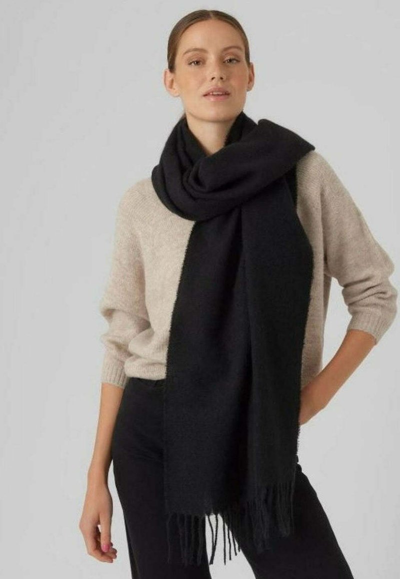 Svart skjerf laget av mykt materiale, med frynser i kantene. Båret løst over en beige genser, tilfører tekstur og kontrast til antrekket.