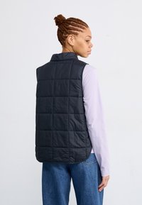 Zwarte gewatteerde vest met een staande kraag, lichtgewicht ontwerp en een zachte textuur, laag over een lichtpaarse lange mouwen top.