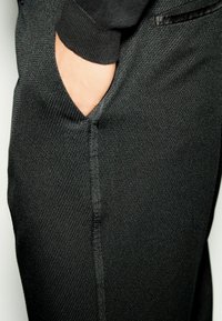 Schwarze Jogginghosen aus strukturiertem Stoff, mit einer seitlichen Tasche und einem glatten, kontrastierenden Bündchen. Sichtbare Nähte entlang der Naht.