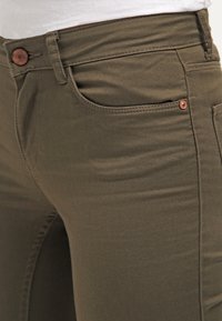 Pantalones ajustados de color verde oliva, confeccionados en una mezcla de algodón elástico. Presentan bolsillos frontales y un botón de tono cobre en la cintura.