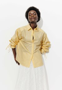 Chemise à boutons rayée jaune avec des manches longues, dotée de liens aux poignets et d'une coupe décontractée. Portée sur une jupe en dentelle blanche.