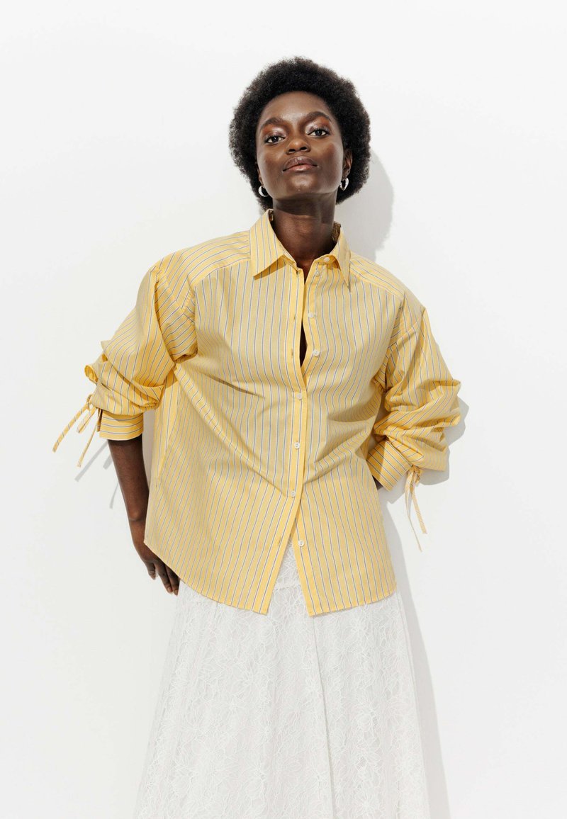 Chemise à boutons rayée jaune avec des manches longues, dotée de liens aux poignets et d'une coupe décontractée. Portée sur une jupe en dentelle blanche.