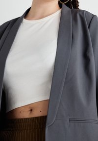 Šedý přizpůsobený blejzr s hladkým povrchem, nošený přes bílý žebrovaný crop top. Outfit obsahuje vysokým pasem hnědý pruhovaný pás.