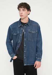 Πουκάμισο από τζιν σε σκούρο μπλε χρώμα, με κουμπιά τύπου snap, δύο τσέπες στο στήθος και κλασικό γιακά. Μακριά μανίκια με μανσέτες που κλείνουν με κουμπιά. Μαλακή υφή.