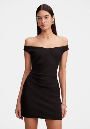 Femme aux cheveux bruns et raides porte une robe noire ajustée, épaules dénudées, des boucles d'oreilles créoles en or et un bracelet en or au poignet droit.