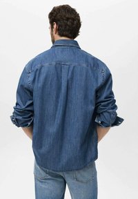 Camicia di jeans con maniche lunghe, colore azzurro chiaro, polsini abbottonati e dettaglio sul retro. Abbinata a jeans blu a contrasto.