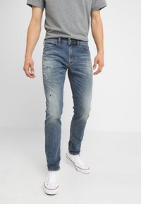 Blå slitna denimjeans med slim fit, med urtvättade sektioner och rivna detaljer, i kombination med en grå t-shirt och vita sneakers.