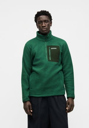 Homme portant une veste polaire Columbia verte avec une poche poitrine zippée, debout devant un fond blanc uni.