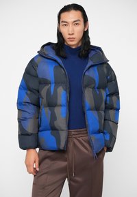 Jachetă puffer în model de camuflaj albastru și gri, cu glugă, închidere cu fermoar și textură matlasată. Modelul poartă un pulover bleumarin dedesubt.