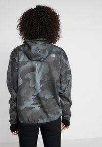Chaqueta ligera con capucha en patrón de camuflaje negro y gris, con puños elásticos y un logo en la parte posterior. Tejido texturizado con un diseño deportivo.