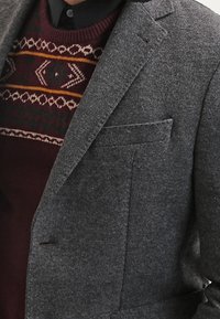 Blazer gris sur mesure avec un revers cranté, porté sur un pull bordeaux présentant un motif géométrique tricoté avec des accents verts et jaunes.