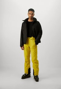 Férfi fekete dzsekiben, fekete ingben, élénk sárga hó nadrágban, amelyhez kerültek a függők, és fekete síbakancsban áll egy fehér háttér előtt.