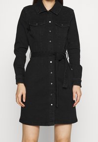 Svart denimskjortklänning med långa ärmar, knäppning fram, två bröstfickor och ett knytbälte i midjan för justerbar passform.