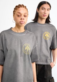 Camisetas gris de gran tamaño con cuello redondo, que presentan un logo circular amarillo en la parte frontal y mangas holgadas.