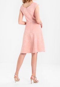 Kvinna klädd i en knälång, ljusrosa mönstrad klänning med korta ärmar och beige högklackade sandaler, vänd bort från kameran.