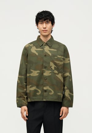 Jeune homme portant une veste camouflage verte et marron avec des boutons et une poche sur la poitrine, debout devant un fond clair uni.