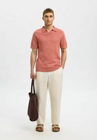 Kortermet, rosa strikket polo med teksturert mønster, kombinert med lyse linbukser og en brun totebag. Sandaler fullfører antrekket.