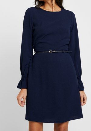 Femme portant une robe bleu marine à manches longues avec des poignets froncés et une fine ceinture noire à la taille, debout devant un fond uni.