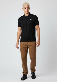 Černé polo tričko s jemným vzorem, krátké rukávy a límeček; spárováno s béžovými kalhotami a černými botami. Čtvercové logo na levé hrudi.