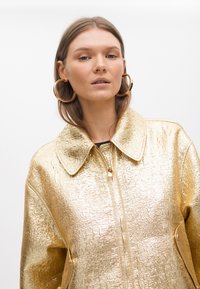 Zlatna metalna jakna na zatvaranje s teksturiranim završetkom, oversize ovratnikom i bočnim džepovima. Model nosi velike okrugle naušnice.