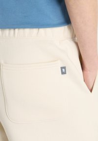 Un pantalon de survêtement beige avec une simple poche arrière et une petite étiquette logo sombre, associé à une chemise bleue, montrant une coupe décontractée et une texture lisse.