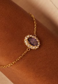 Gouden armband met een centrale ovale paarse steen omringd door heldere rhinestones; heeft een fijne ketting en verfijnde details.