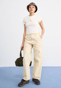 Witte geribbelde crop top met gele geschulpte randen en bloemetjesmotief, gecombineerd met beige cargobroek en sneakers met donkere zolen, terwijl ze een groene tas vasthoudt.
