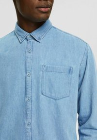 Camisa de mezclilla azul claro con cierre frontal de botones, cuello puntiagudo, un bolsillo en el pecho y botones contrastantes. Textura suave y diseño ajustado.