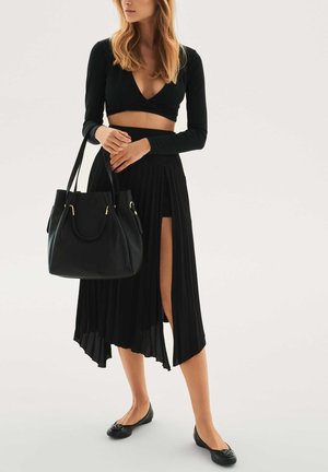Black pleated skirt with a high slit paired with a black crop top, flat black shoes, and a black handbag with gold hardware accents.