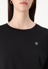 Black cotton t-shirt with a round neckline and short sleeves. Features a small grey logo on the left chest. Smooth texture.