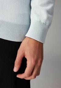 Lyseblå strikket sweater med ribbet kant, close-up af en hånd hvile mod et mørkt stof, der viser tekstur og farvekontrast.