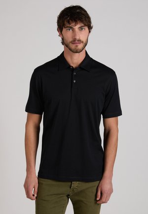 Mann mit Bart, der ein schwarzes Poloshirt mit kurzen Ärmeln und olivgrüne Hosen trägt, vor einem einfarbigen hellgrauen Hintergrund stehend.