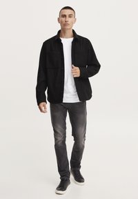 Camisa negra de botones con dos bolsillos en el pecho, llevada sobre una camiseta blanca. Combinada con jeans grises y zapatillas negras. Textura de tela suave.