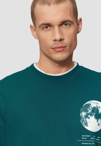 Türkise Sweatshirt mit geripptem Rundhalsausschnitt, auf der linken Seite mit einem weißen Mondgrafik und Textdruck. Weiches, strukturiertes Material.